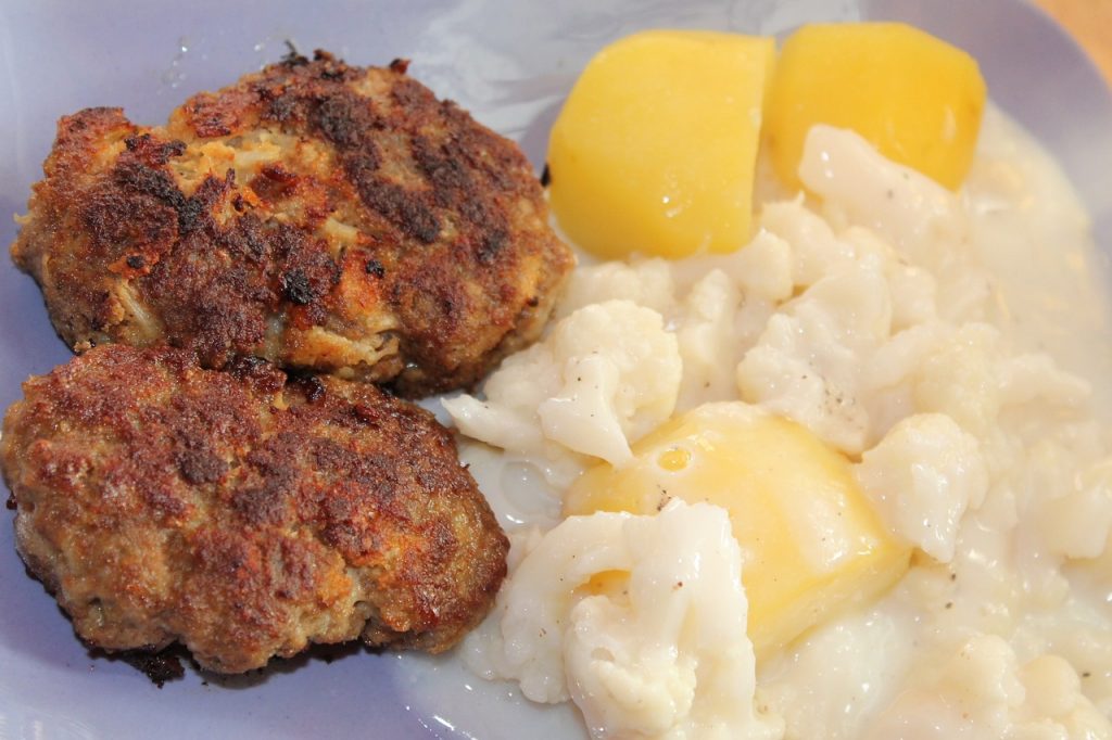 Zwei goldbraune Frikadellen mit Kartoffeln und Blumenkohl auf einem Teller, typisch für deutsche Küche.