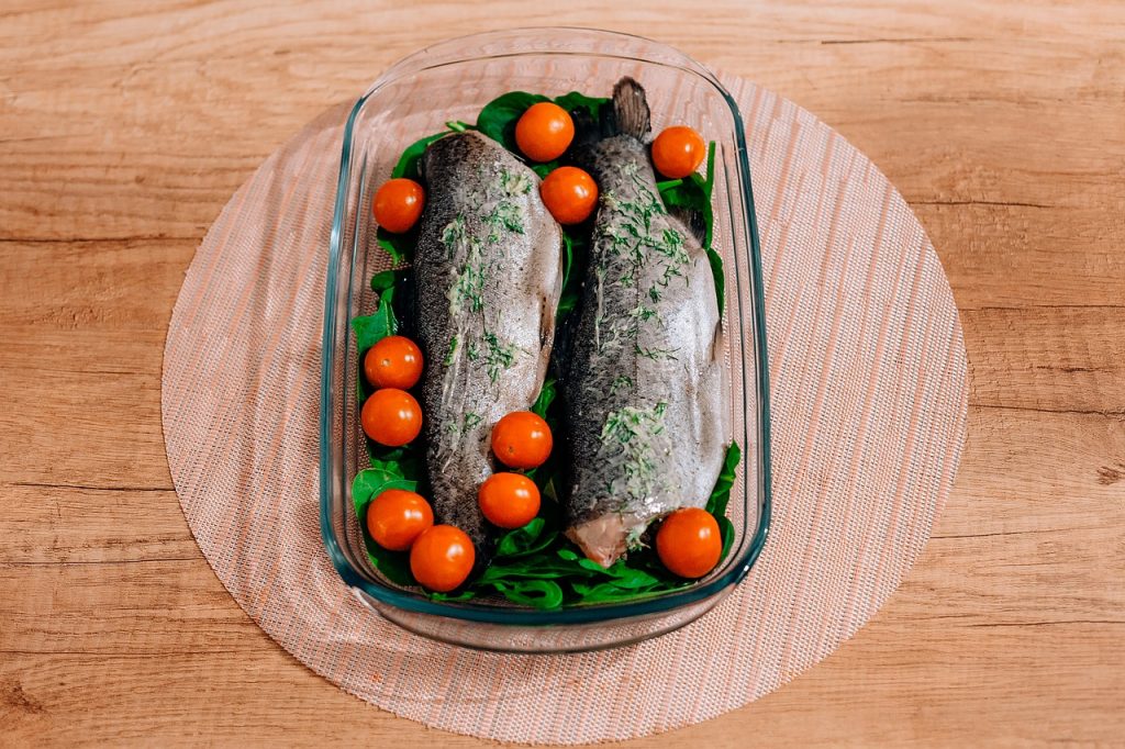 Zwei frische Fische auf einem Bett aus Spinat und umgeben von Kirschtomaten in einer Glasform.