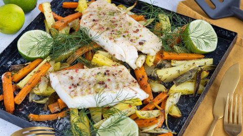 Gebackener Fischfilet auf einem Blech mit buntem Gemüse, darunter Karotten und Kartoffeln, garniert mit Limetten und Dill.