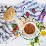 Eine Schüssel Gazpacho mit frischen Kräutern, umgeben von Brot, Kirschtomaten und Olivenöl auf einem gestreiften Tuch.