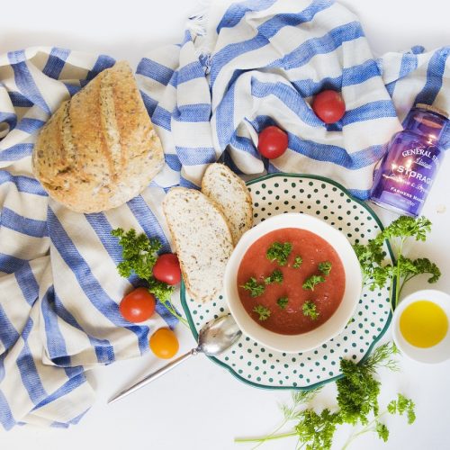 Eine Schüssel Gazpacho mit frischen Kräutern, umgeben von Brot, Kirschtomaten und Olivenöl auf einem gestreiften Tuch.