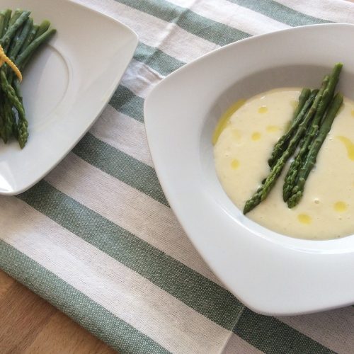 Frischee Spargelsuppe auf einem Teller mit einer Beilage von Spargelstangen, serviert auf einem gestreiften Tischläufer.