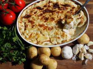 Ein köstliches Kartoffelgratin mit goldbrauner Kruste, umgeben von frischen Tomaten, Kartoffeln, Knoblauch und Petersilie.