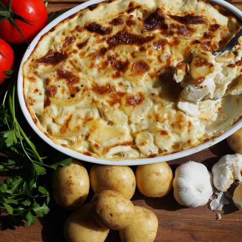 Ein köstliches Kartoffelgratin mit goldbrauner Kruste, umgeben von frischen Tomaten, Kartoffeln, Knoblauch und Petersilie.