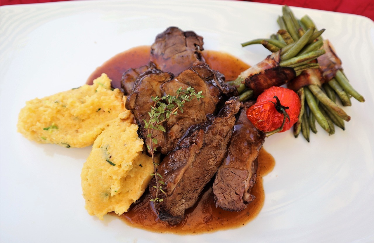 Ein köstliches Rindfleischgericht mit Polenta, grünen Bohnen und einer Tomate, serviert auf einem weißen Teller.