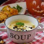 Schüssel mit Blumenkohlsuppe, garniert mit Croutons und Petersilie, auf einem karierten Tisch. Herbstliche Dekoration im Hintergrund.