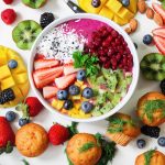 Bunte Smoothie-Bowl mit Erdbeeren, Heidelbeeren, Kiwi, Mango und Kokosnuss, umgeben von frischen Beeren und Muffins.