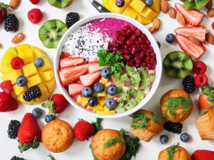Bunte Smoothie-Bowl mit Erdbeeren, Heidelbeeren, Kiwi, Mango und Kokosnuss, umgeben von frischen Beeren und Muffins.