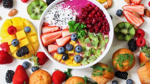 Bunte Smoothie-Bowl mit Erdbeeren, Heidelbeeren, Kiwi, Mango und Kokosnuss, umgeben von frischen Beeren und Muffins.