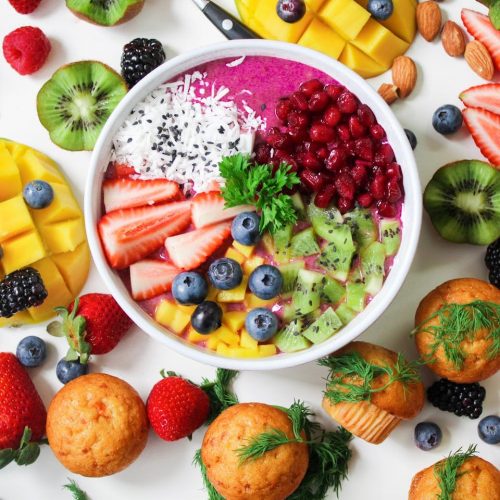 Bunte Smoothie-Bowl mit Erdbeeren, Heidelbeeren, Kiwi, Mango und Kokosnuss, umgeben von frischen Beeren und Muffins.