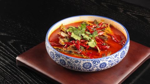 Schüssel mit scharfer Paprika-Suppe, gefüllt mit getrockneten Chilis und frischen Kräutern, auf einem Holzuntersetzer.