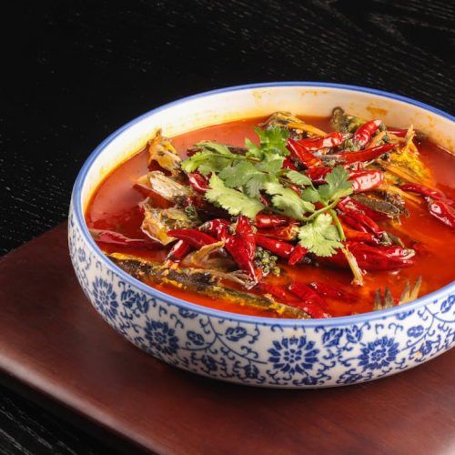 Schüssel mit scharfer Paprika-Suppe, gefüllt mit getrockneten Chilis und frischen Kräutern, auf einem Holzuntersetzer.