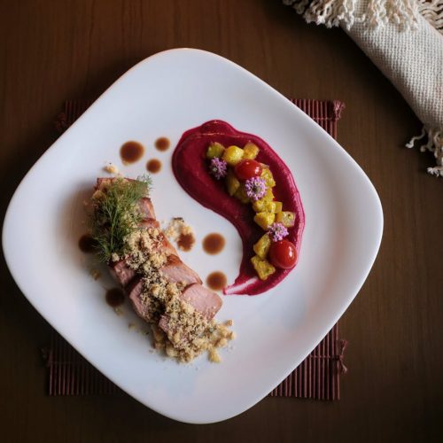 Ein kunstvoll angerichtetes Gourmetgericht mit Fleisch, Beerenpüree und bunten Beilagen auf einem weißen Teller.