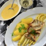 Ein Teller mit gebratenem Fisch, Spargel, Kartoffeln und einer gelben Sauce, serviert mit Zitronenscheiben und einem Glas Weißwein.