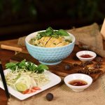 Schüssel mit asiatischer Nudelsuppe, frischem Gemüse, Kräutern und Soßen auf einem Holztisch.