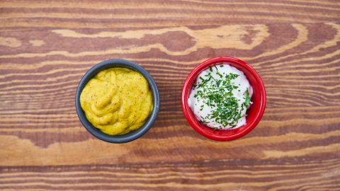 Eine Schüssel mit Senf und eine Schüssel mit Joghurt, garniert mit frischen Kräutern auf einem Holzuntergrund.