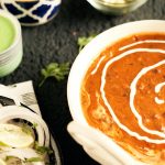 Ein Teller mit indischem Gericht in Soße, garniert mit Sahne, umgeben von Beilagen wie Zwiebeln und Saucen.