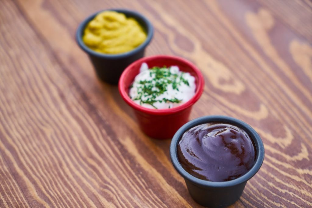 Drei verschiedene Dips in kleinen Schalen: Senf, Joghurt-Dip mit Kräutern und BBQ-Sauce auf einem Holztisch.