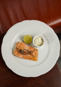 Gegrilltes Lachsfilet auf einem weißen Teller mit Limettenscheibe und einer Portion Sauce, ideal für Feinschmecker und Fischliebhaber.