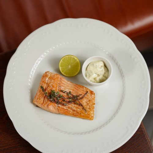 Gegrilltes Lachsfilet auf einem weißen Teller mit Limettenscheibe und einer Portion Sauce, ideal für Feinschmecker und Fischliebhaber.