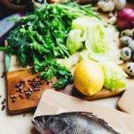 Frischer Kabeljau auf einem Holzbrett, umgeben von Gemüse, Kräutern und einer Zitrone, ideal für gesunde Rezepte.