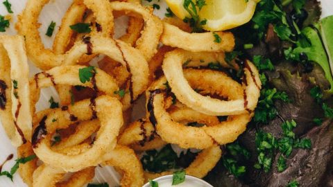 Frittierte Calamari-Ringe mit frischer Zitrone und Kräutern, serviert mit einer Dip-Sauce auf einem weißen Teller.