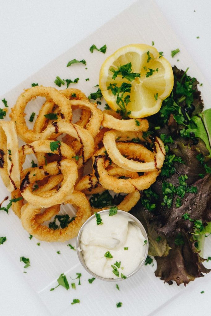 Frittierte Calamari-Ringe mit frischer Zitrone und Kräutern, serviert mit einer Dip-Sauce auf einem weißen Teller.