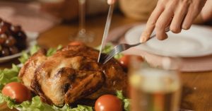 Geröstetes Hähnchen auf einem Tisch, umgeben von frischem Gemüse und Tomaten, bereit zum Servieren, mit Besteck zum Schneiden.