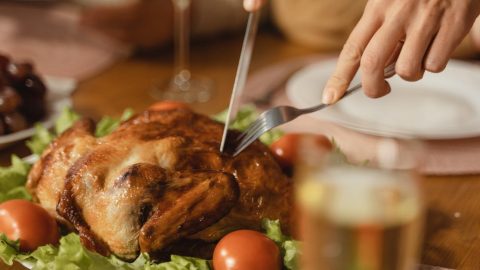Geröstetes Hähnchen auf einem Tisch, umgeben von frischem Gemüse und Tomaten, bereit zum Servieren, mit Besteck zum Schneiden.