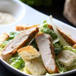 Frischer Hähnchensalat mit Caesar-Dressing, Croutons und grünen Blättern auf einem weißen Teller.