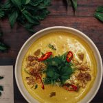 Eine cremige Humus-Suppe mit frischen Kräutern, roten Chilischoten und knusprigen Zwiebelstücken in einer Schüssel.