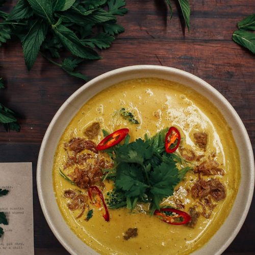 Eine cremige Humus-Suppe mit frischen Kräutern, roten Chilischoten und knusprigen Zwiebelstücken in einer Schüssel.