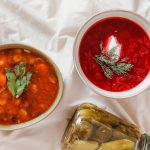 Zwei Schalen mit hausgemachten Suppen, eine mit Gemüse und die andere mit Rote-Bete, begleitet von Brot und eingelegten Gurken.