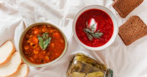Zwei Schalen mit hausgemachten Suppen, eine mit Gemüse und die andere mit Rote-Bete, begleitet von Brot und eingelegten Gurken.