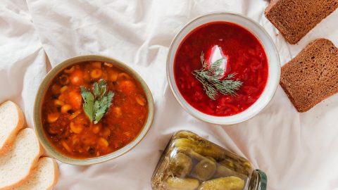 Zwei Schalen mit hausgemachten Suppen, eine mit Gemüse und die andere mit Rote-Bete, begleitet von Brot und eingelegten Gurken.