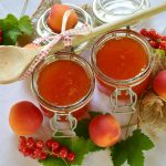 Zwei Gläser mit hausgemachter Marmelade, umgeben von frischen Aprikosen und roten Johannisbeeren, mit einem Holzlöffel.