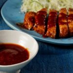 Panierte Hähnchenbrustscheiben auf einem blauen Teller mit einer Schüssel Sauce, serviert mit Salatbeilage.