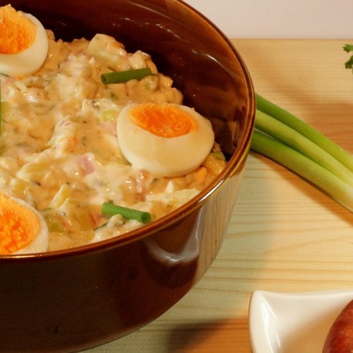 Kartoffelsalat mit hartgekochten Eiern, Wurst, Radieschen und Frühlingszwiebeln in einer braunen Schüssel.