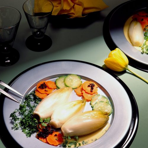 Gekochter Chicorée auf einem Teller mit Karotten, Zucchini und einer Sauce, serviert mit Gläsern und Dekoration.