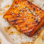 Geräucherter und gegrillter Lachs auf einer Reisbasis mit Sesam bestreut, serviert in einer umweltfreundlichen Verpackung.