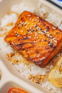 Geräucherter und gegrillter Lachs auf einer Reisbasis mit Sesam bestreut, serviert in einer umweltfreundlichen Verpackung.