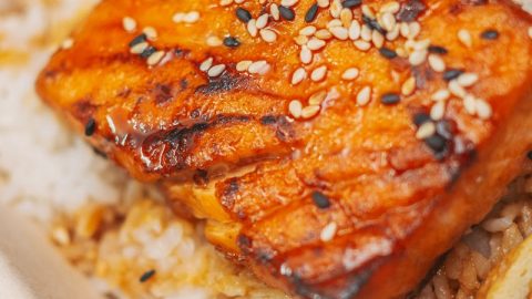 Geräucherter und gegrillter Lachs auf einer Reisbasis mit Sesam bestreut, serviert in einer umweltfreundlichen Verpackung.