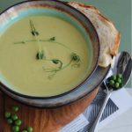 Schüssel mit cremiger Erbsen-Minze-Suppe, garniert mit Erbsen und frischen Kräutern, serviert mit Brot auf einem Holzbrett.