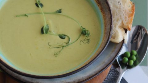 Schüssel mit cremiger Erbsen-Minze-Suppe, garniert mit Erbsen und frischen Kräutern, serviert mit Brot auf einem Holzbrett.