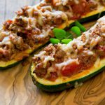 Gefüllte Zucchinihälften mit Hackfleisch, Tomaten und geschmolzenem Käse, serviert auf einem Holzbrett.