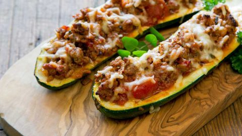 Gefüllte Zucchinihälften mit Hackfleisch, Tomaten und geschmolzenem Käse, serviert auf einem Holzbrett.