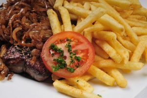 Ein saftiges Steak mit karamellisierten Zwiebeln, serviert mit knusprigen Pommes Frites und einer Scheibe Tomate mit Petersilie.