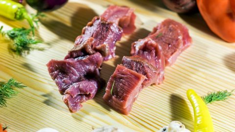 Zwei Stücke frisches Fleisch auf einem Holzbrett, umgeben von Gemüse wie Paprika, Pilzen und Kräutern.