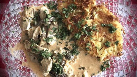 Ein Teller mit zarten Fleischstücken in Sahnesauce und knusprigen Kartoffelpuffern, garniert mit frischen Kräutern.