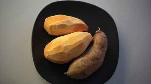 Drei Süßkartoffeln, zwei geschälte und eine ungeschälte, auf einem schwarzen Teller präsentiert, ideal für Rezepte und Ernährung.